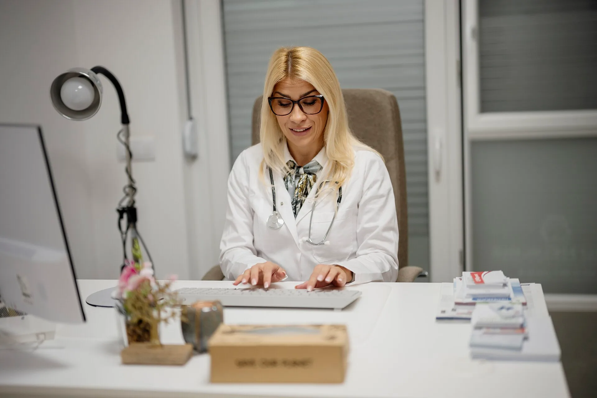 A doctor sits at her desk in a medical office. She is wearing a white coat and glasses. She uses a computer and takes notes while looking engaged. When you are the authority: earning deference without a costume